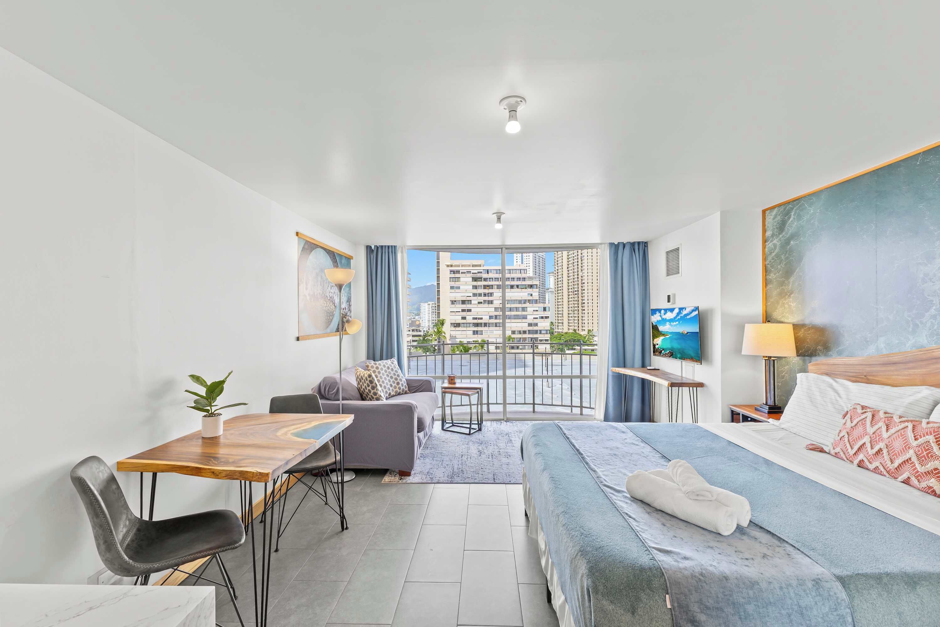 A hotel room with a bed, a desk, a chair, a sofa, a TV, and a balcony with a city view.