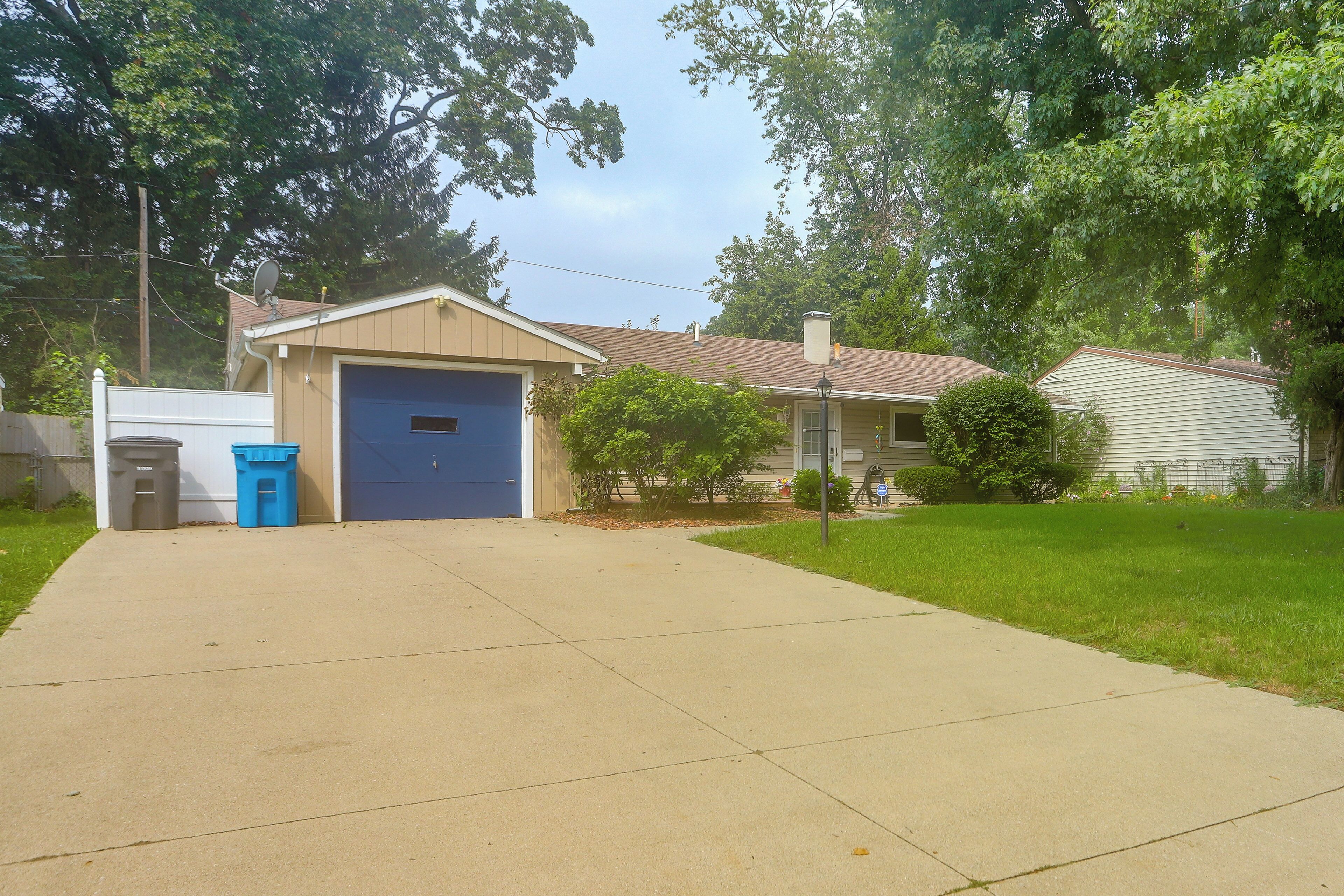 A residential driveway leading to a garage with a blue door, a satellite dish, and a trash bin.