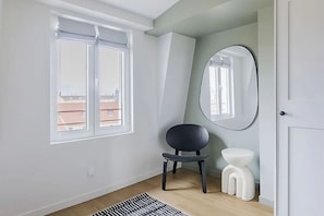 A room with a large window, a mirror, a chair, a small stool, and a patterned rug. - Elegant 3-Bedroom Paris 6th Apartment with Eiffel Tower Views & A/C (Paris)