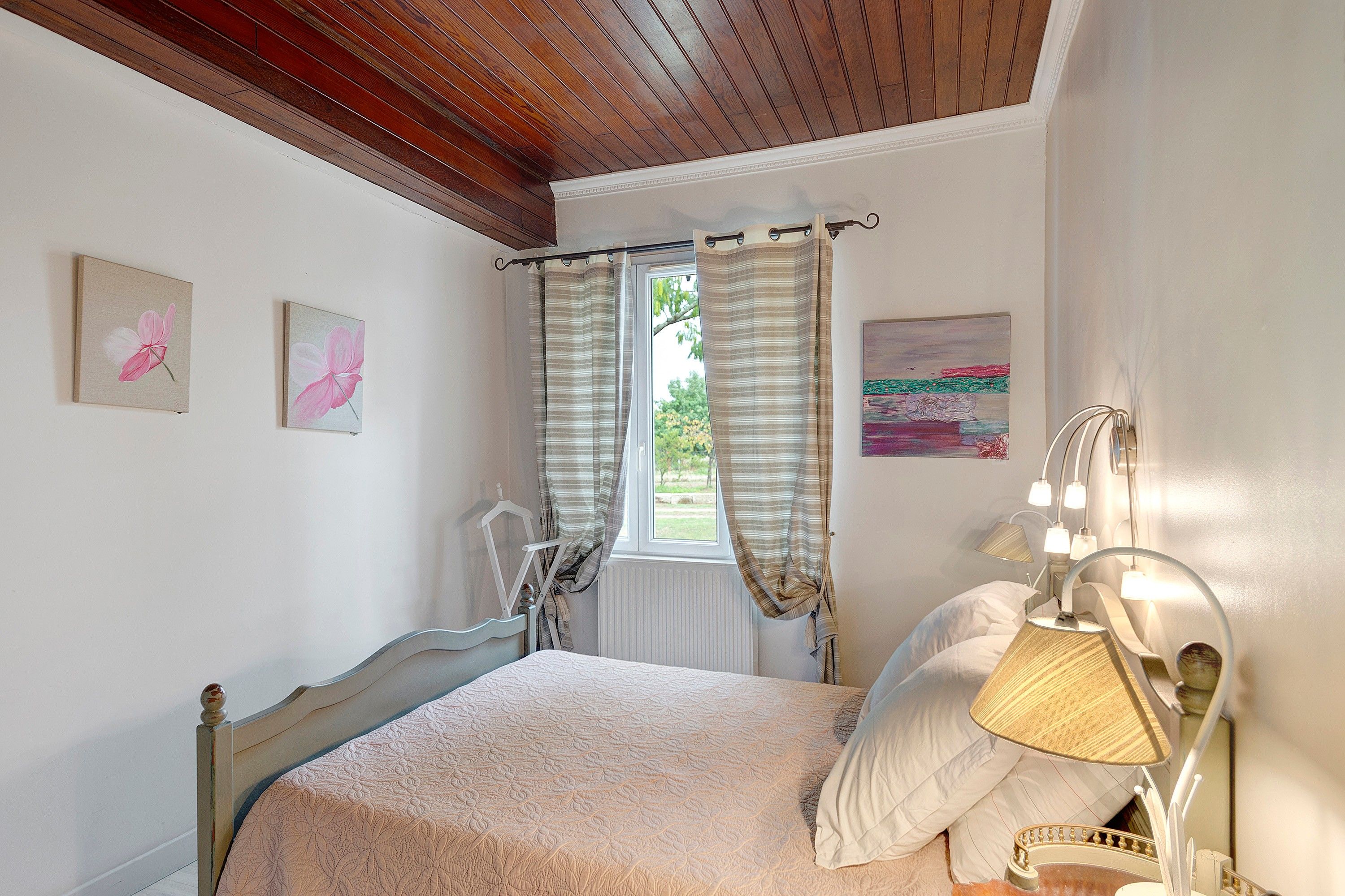 Une chambre à coucher avec un lit, une fenêtre avec des rideaux, une lampe de chevet et deux tableaux accrochés au mur.