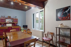 A dining area with a wooden table, chairs, a radiator, a window, and a shelf with items. - Gîte de Bossuet – Wi-Fi, Garden & Air Conditioning near Bordeaux & Atlantic Coast (Saint-Denis-de-Pile)