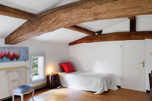 A bedroom with a wooden ceiling, a bed with a white bedspread and a red pillow, a nightstand with a lamp, a chair, and a painting on the wall. - Gîte de Bossuet – Wi-Fi, Garden & Air Conditioning near Bordeaux & Atlantic Coast (Saint-Denis-de-Pile)