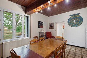 A dining area with a wooden table and chairs, a window with a view of greenery, and a wall with a clock and artwork. - Gîte de Bossuet – Wi-Fi, Garden & Air Conditioning near Bordeaux & Atlantic Coast (Saint-Denis-de-Pile)