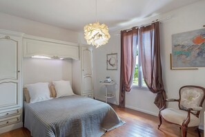 A bedroom with a bed, a chair, a hanging light fixture, a window with curtains, and a painting on the wall. - Gîte de Bossuet – Wi-Fi, Garden & Air Conditioning near Bordeaux & Atlantic Coast (Saint-Denis-de-Pile)