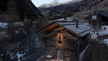 En stuga byggd av trä med ett sluttande tak, en stig av sten som leder till ingången och en utsikt över en flod och berg i bakgrunden.
