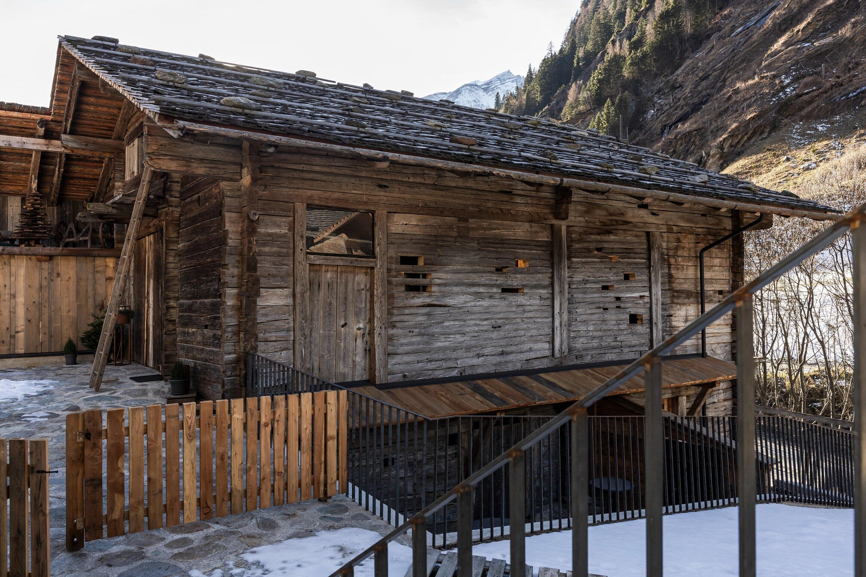 Una cabaña de madera rústica con techo pronunciado, rodeada de nieve y montañas.
