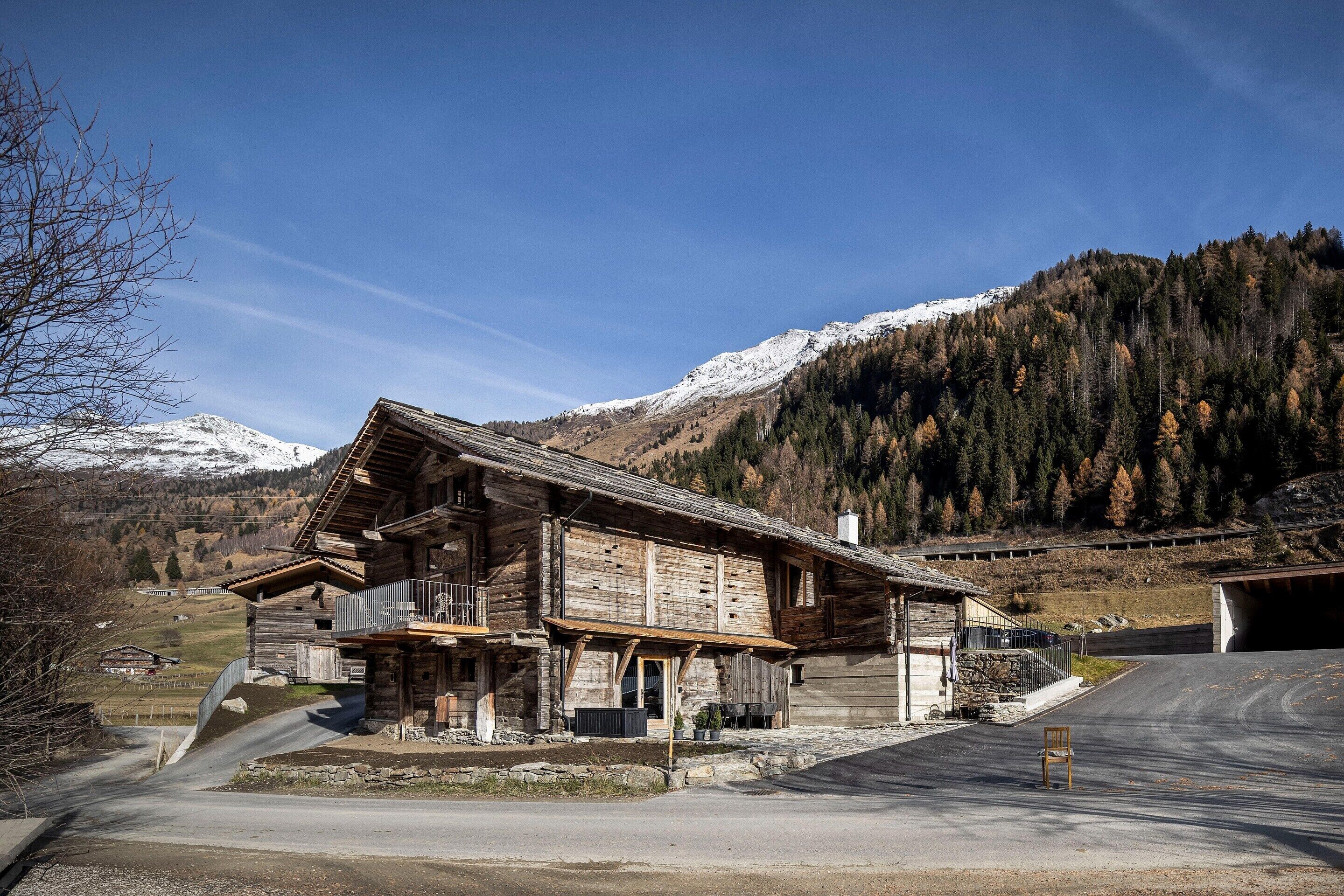 A rustic wooden building with a balcony, set against a backdrop of snow-capped mountains and a clear sky.