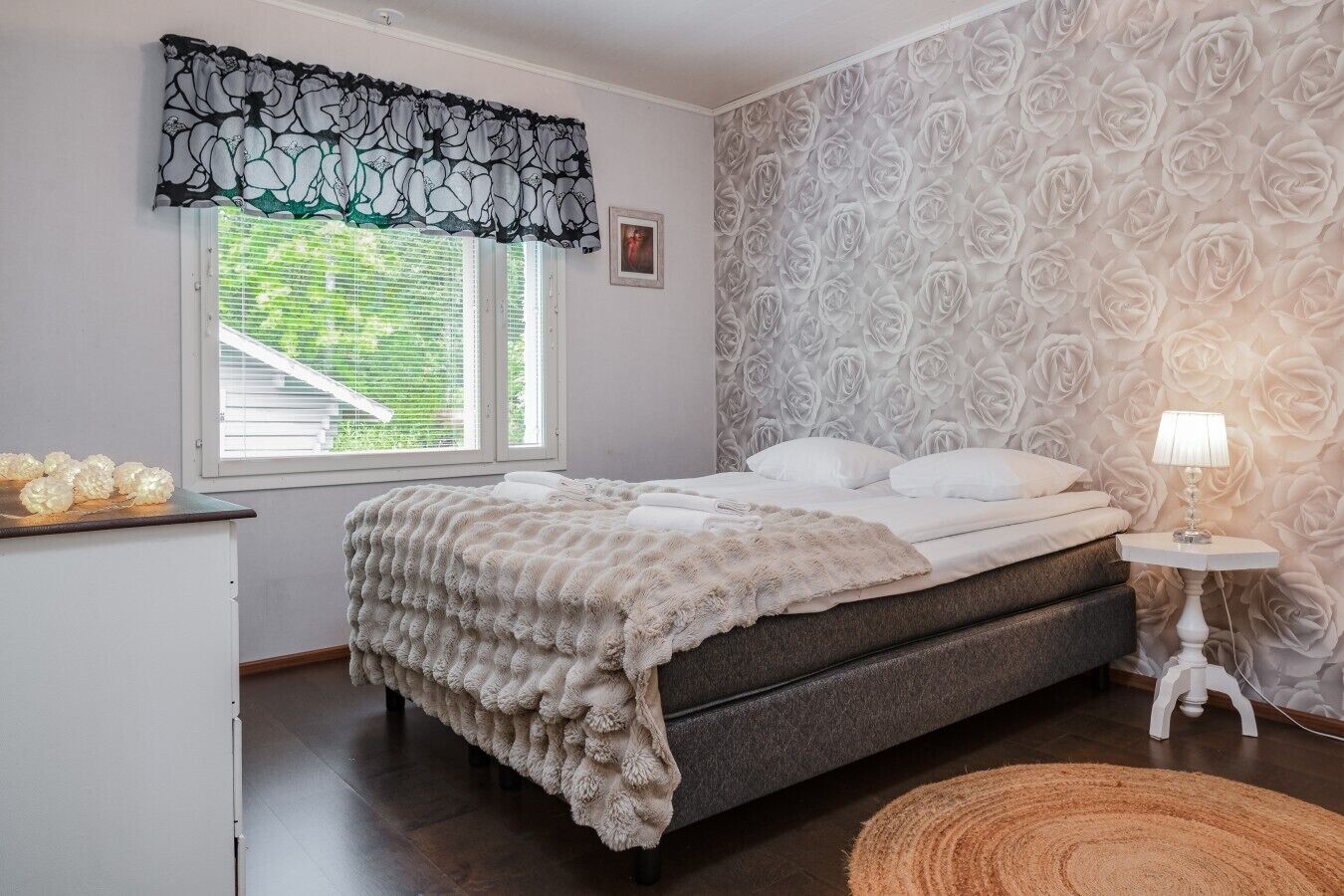 Une chambre à coucher avec un lit, une table de chevet avec une lampe, une fenêtre avec des stores et un mur orné d’un motif floral.