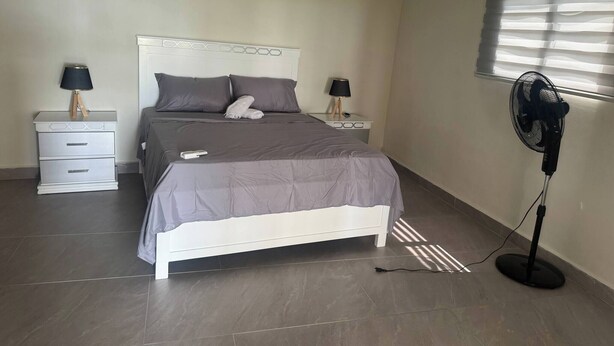A bedroom with a bed, bedside tables, lamps, a standing fan, and a window with blinds. - Rico Puerto magic spot oeste (Cabrera)