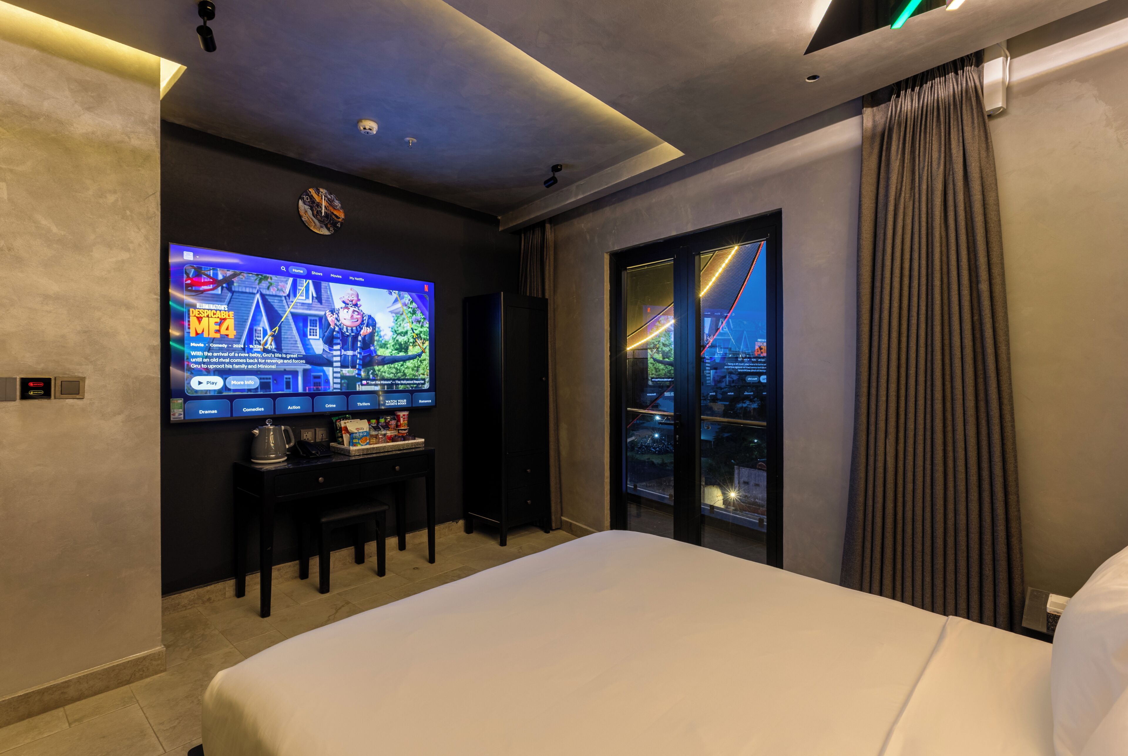 A hotel room with a large flat-screen TV displaying a movie, a bed, a coffee table with snacks, and a view of a cityscape through a glass door.