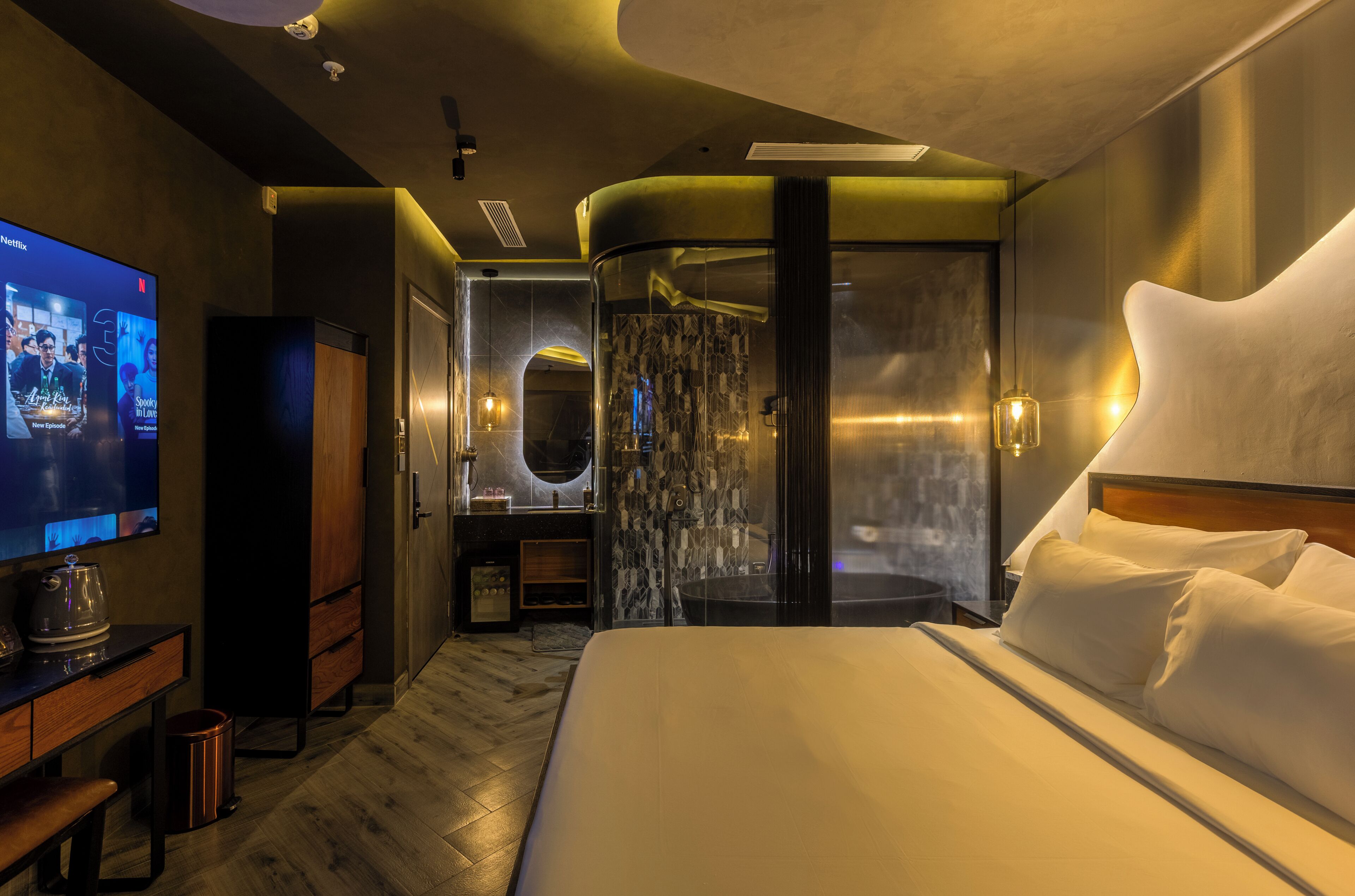 A modern hotel room with a large bed, a TV, a minibar, and a bathroom with a glass shower enclosure.