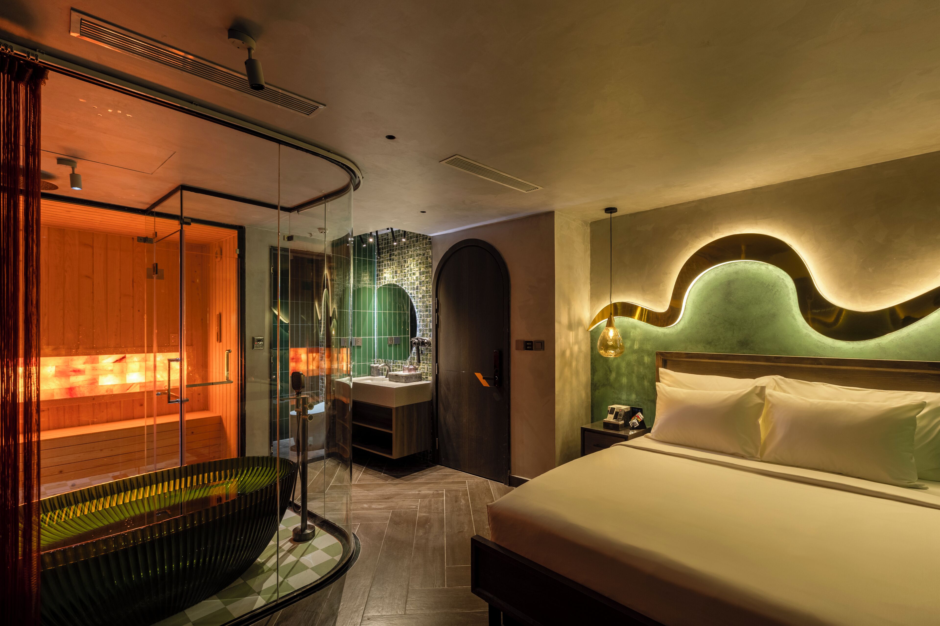 A modern hotel room with a large bed, a glass-enclosed sauna, and a separate bathroom with a green-tiled wall.