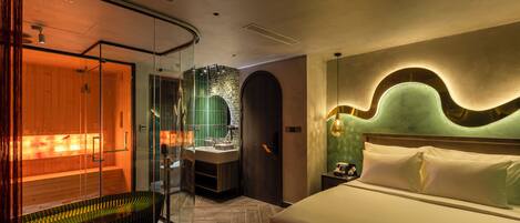 A modern hotel room with a large bed, a glass-enclosed sauna, and a separate bathroom with a green-tiled wall.