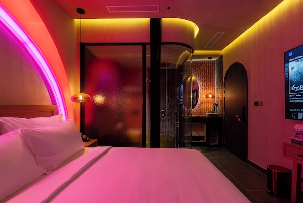 A hotel room with a bed, a nightstand, a wall-mounted TV, and a bathroom visible through a glass partition.
