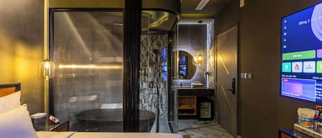 A hotel room with a bed, a desk with a computer, a TV, and a bathroom with a shower.