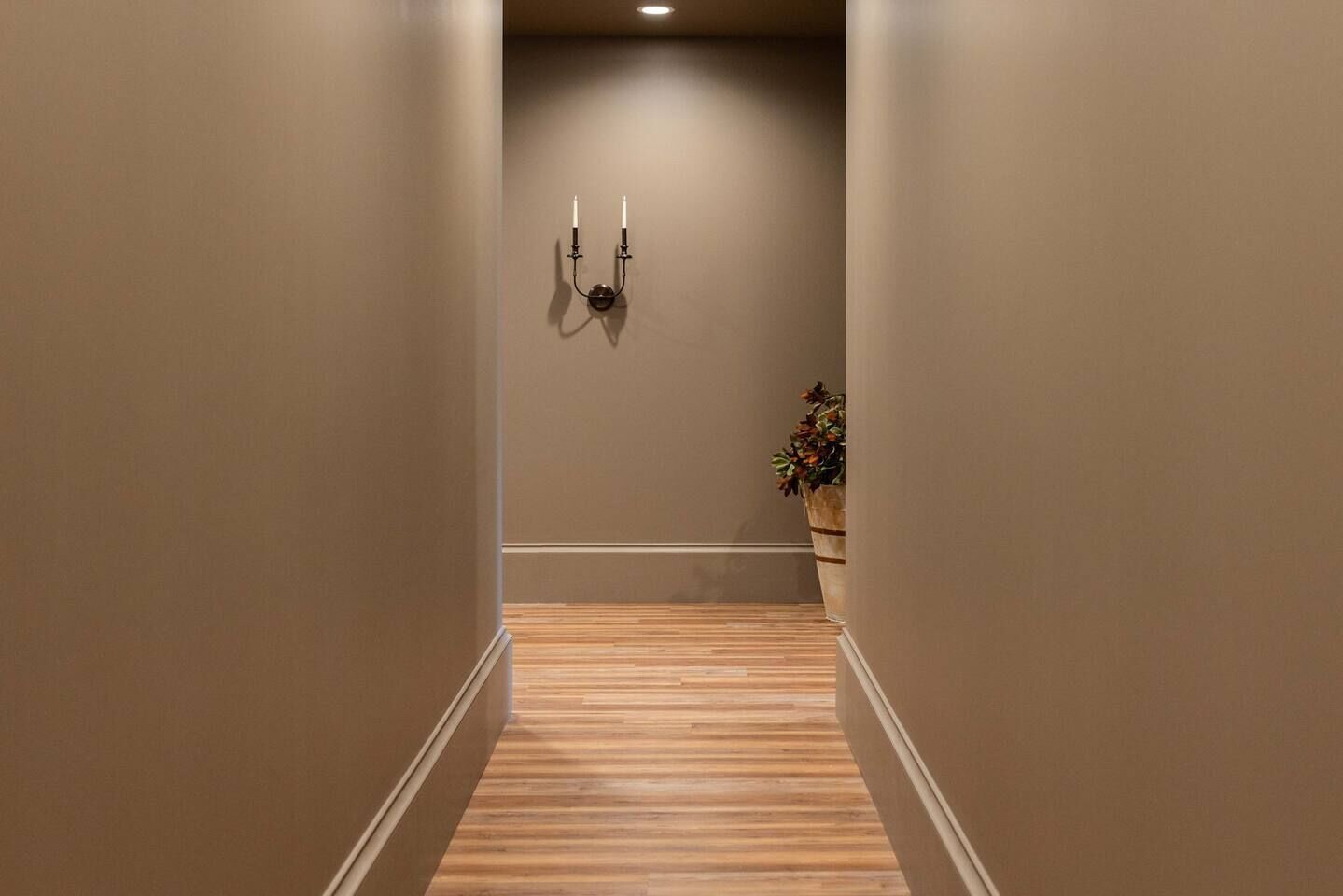 A hallway with wooden flooring, beige walls, and a wall-mounted light fixture.