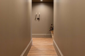 A narrow hallway with wooden flooring and beige walls, leading to a room with a potted plant.