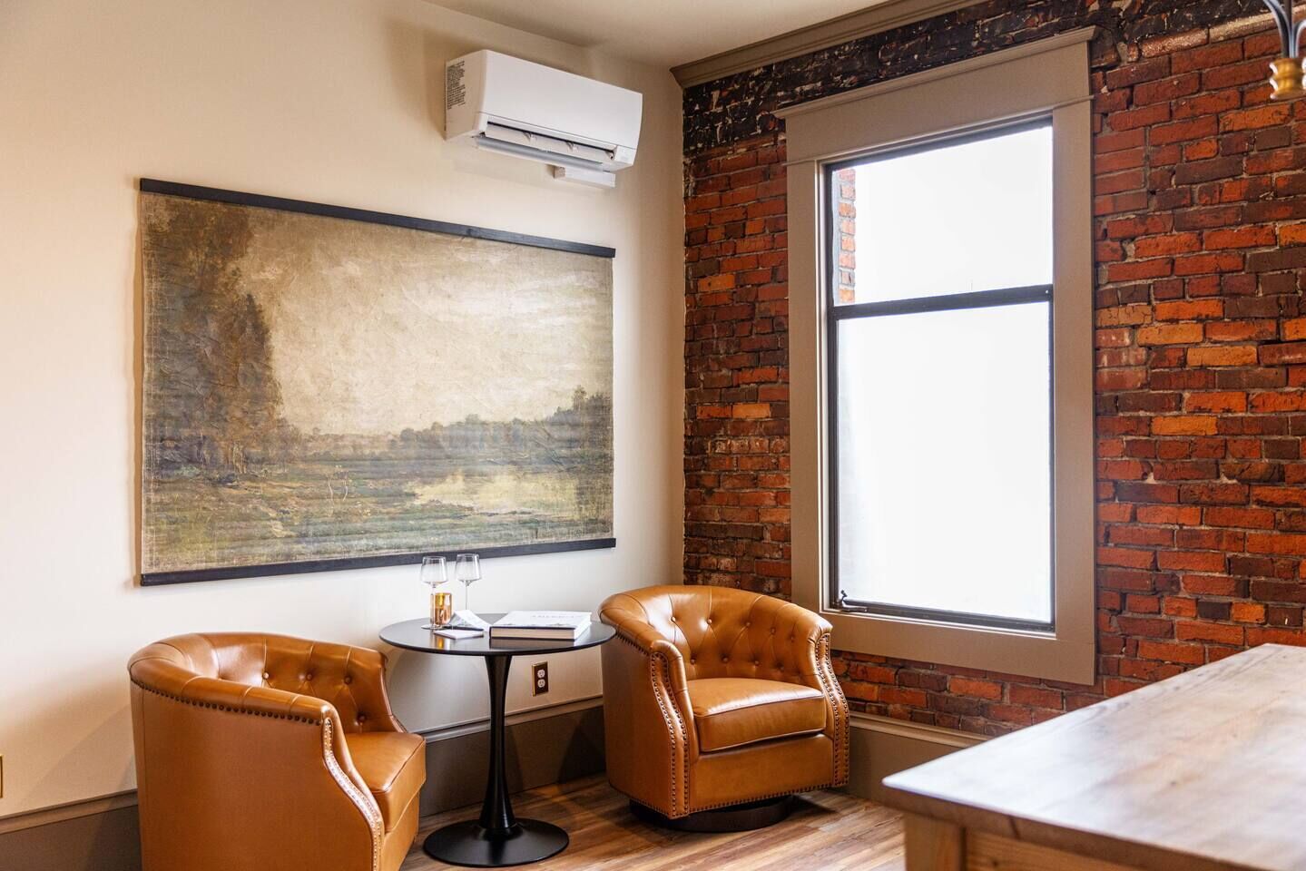 A room with a brick wall, a large window, two leather chairs, a small round table with a book and glasses, and a painting.