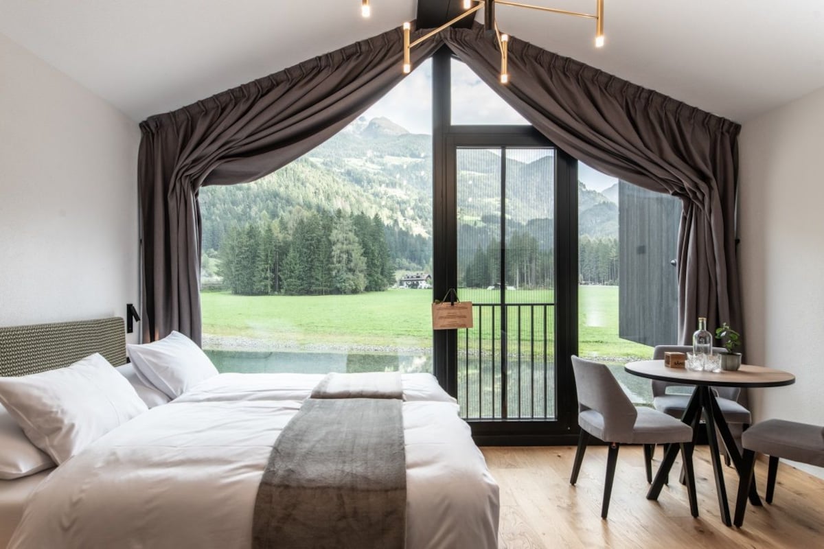 A bedroom with a large bed, a dining table, and a view of a mountainous landscape through a sliding door.