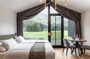 A bedroom with a large bed, a dining table, and a view of a mountainous landscape through a sliding door.