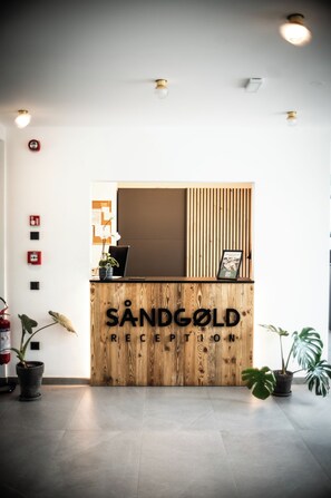 A reception desk with a wooden finish and the name 'SANDGOLD RECEPTION' displayed. There are potted plants on either side of the desk, and a fire alarm is mounted on the wall to the left.
