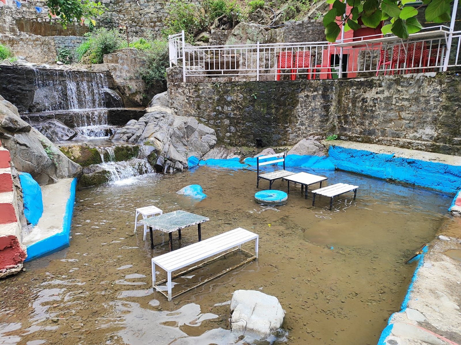 Ein Naturbad mit Bänken und einem kleinen Wasserfall, umgeben von Steinmauern und Vegetation.