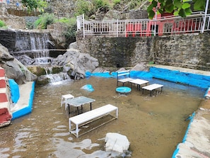 Una piscina natural con bancos y una pequeña cascada, rodeada de muros de piedra y vegetación.