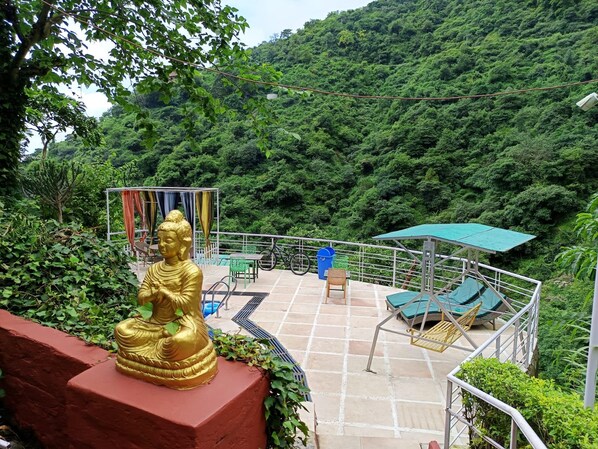 A Buddha statue on a red pedestal, a covered seating area with chairs and a swing, and a view of a lush green hillside.