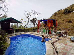 Una piscina con una persona nadando, un área de descanso techada y una ladera al fondo.