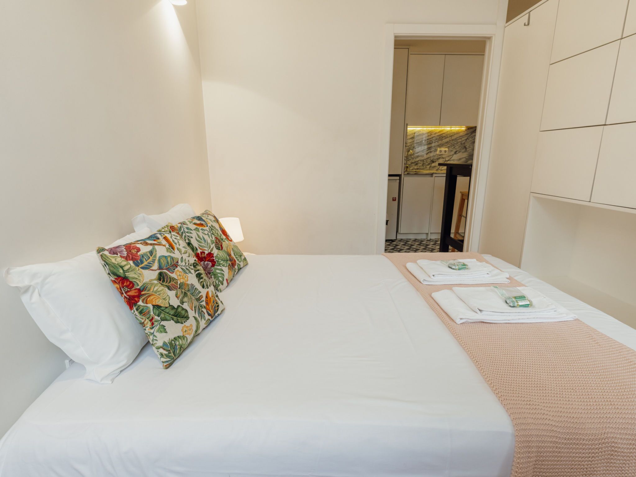 A hotel room with a bed, a pillow, a bedside table, and a view into the kitchen area.