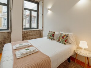 A hotel room with a bed, pillows, a bedside table with a lamp, and a window with a view of buildings. - Apartment  by Interhome (Porto)