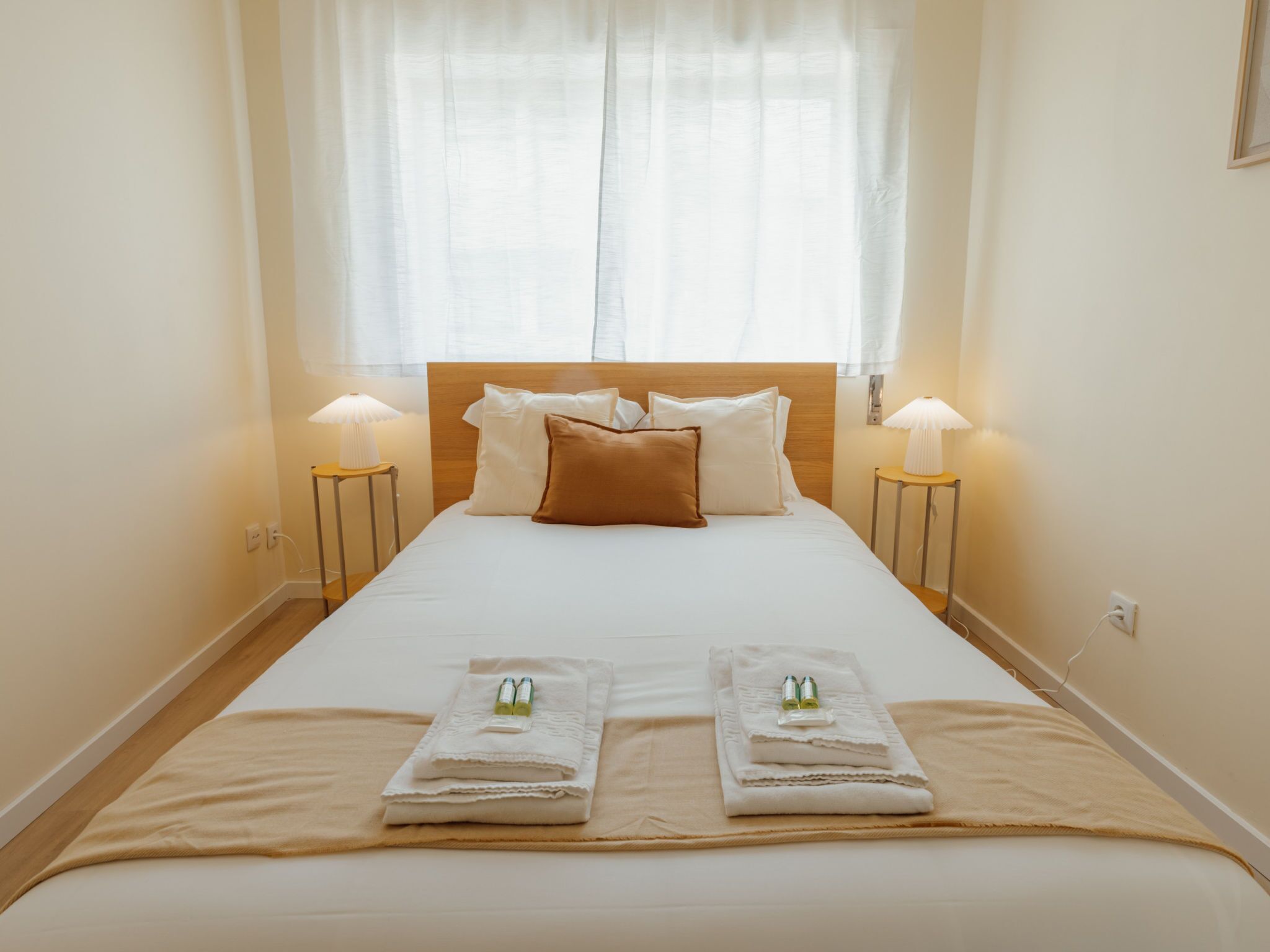 A neatly made bed with a wooden headboard, two bedside lamps, and towels with bottles on the bed.