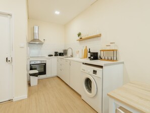 Uma cozinha compacta com armários brancos, forno embutido, máquina de lavar e mesa de madeira.