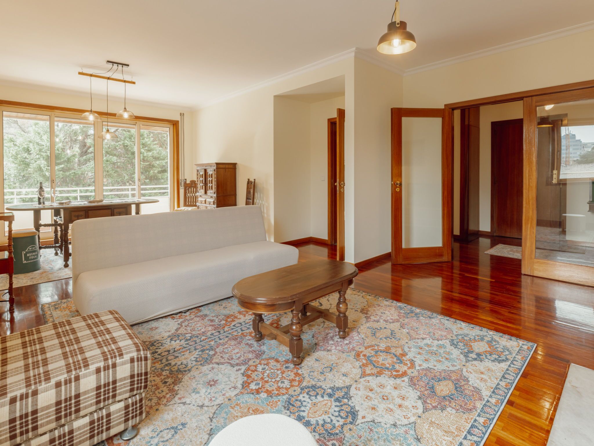 A spacious living room with a sofa, a wooden coffee table, and a patterned rug. There is a dining area with a table and chairs visible through a glass door.