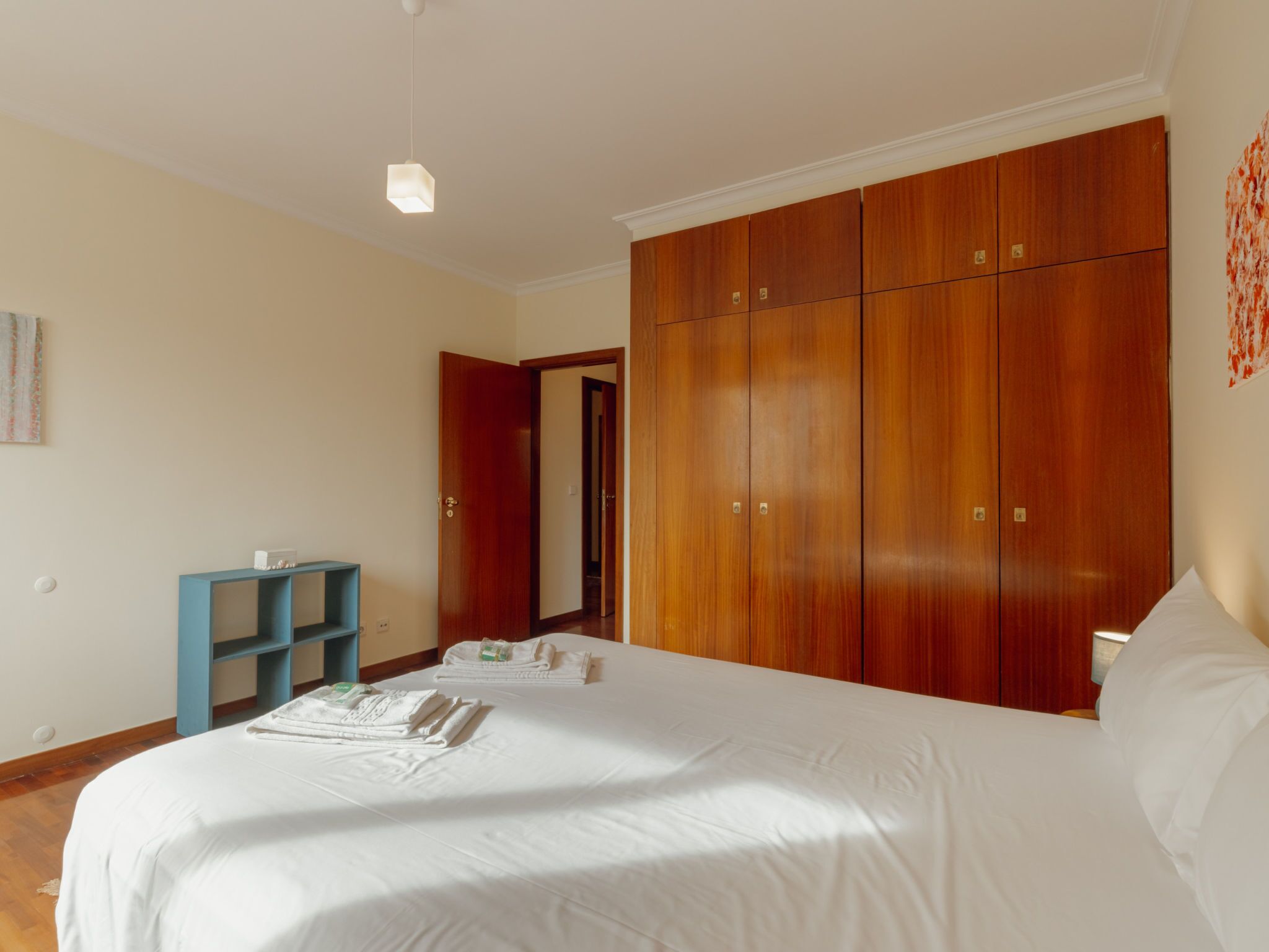 A hotel room with a large bed, a wardrobe, a desk with a chair, and a hanging light fixture.
