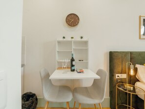 A small, modern room with a white table, two grey chairs, a clock on the wall, a shelf with plants, and a bed with a green headboard. - Apartment  by Interhome (Porto)