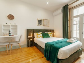 Quarto de hotel com cama, escrivaninha, cadeira, relógio e janela com cortinas.
