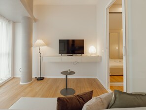 Ein modernes Wohnzimmer mit einem an der weißen Wand befestigten Flachbildfernseher, einer Stehlampe, einem kleinen runden Tisch mit einer Pflanze und einer Couch mit Kissen.
