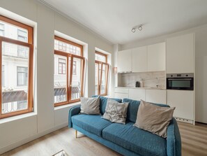 Ett modernt vardagsrum med en blå soffa, ett kök med vita skåp och stora fönster med träramar.