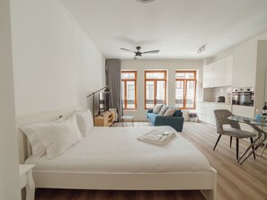 Ein modernes Hotelzimmer mit einem großen Bett, einem Schreibtisch, einem Sessel und Blick auf die Stadt durch große Fenster.