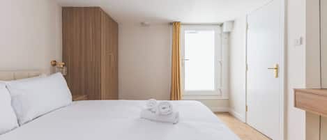 A hotel room with a large bed, a wooden wardrobe, a window with curtains, and a wooden bedside table.