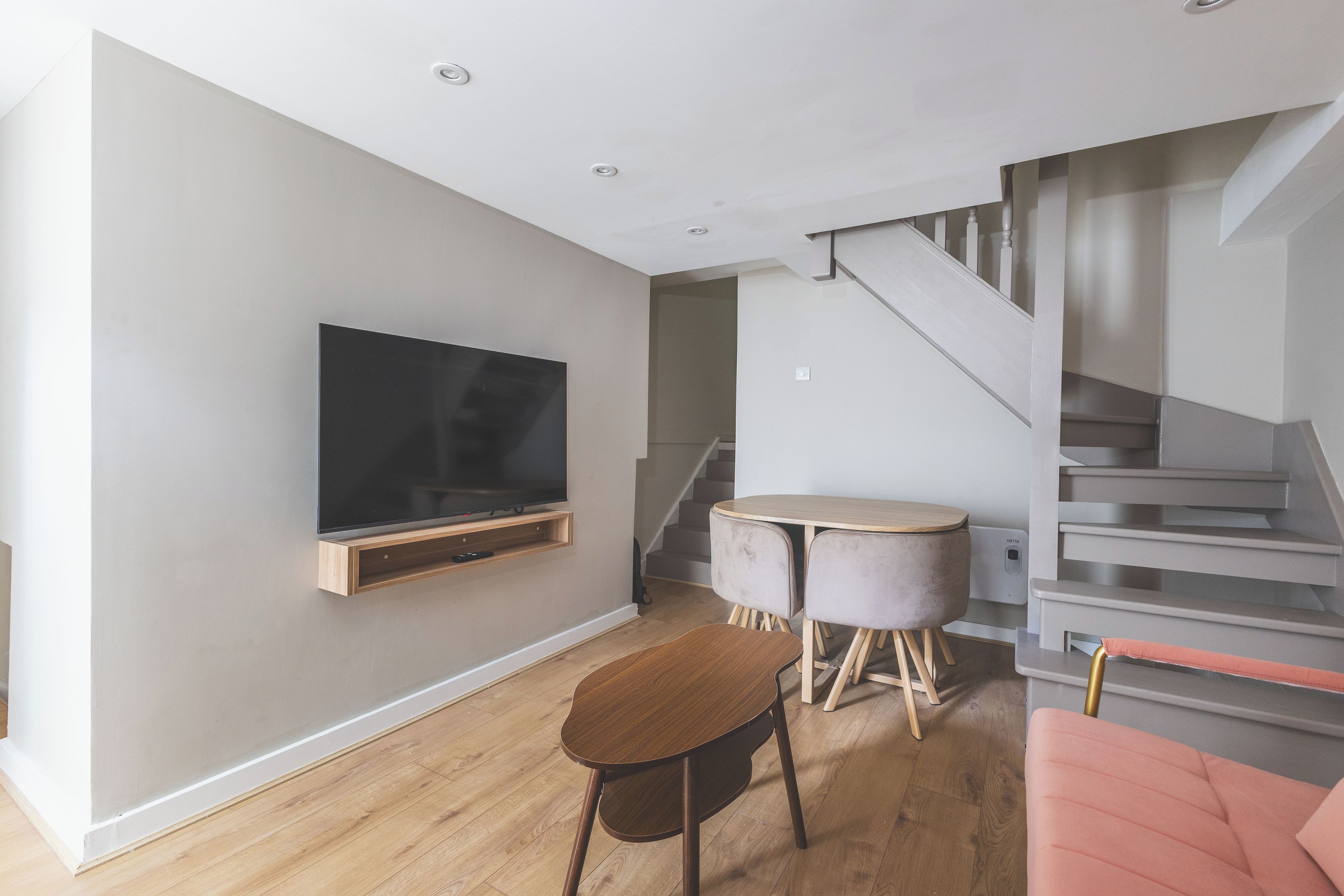 Una sala moderna con televisor de pantalla plana en la pared, una mesa redonda de madera, un sillón rosa y una escalera que conduce a un piso superior.