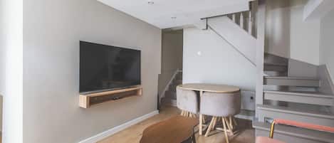 Una sala moderna con televisor de pantalla plana en la pared, una mesa redonda de madera, un sillón rosa y una escalera que conduce a un piso superior.