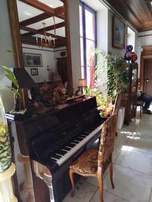 Un piano de cola en una habitación bien iluminada, con un espejo grande, una silla de madera y varios objetos decorativos, incluyendo un reloj y un florero.