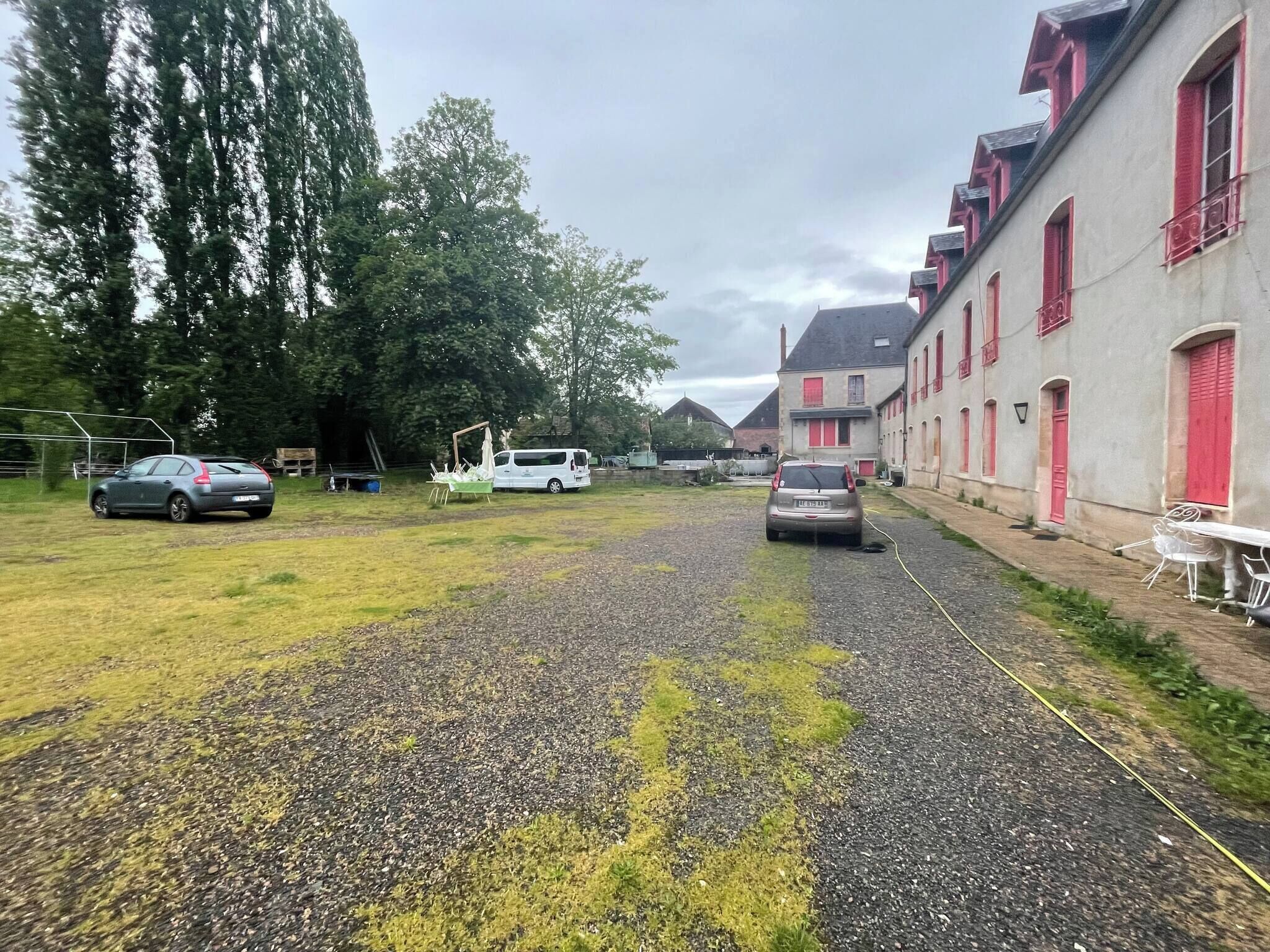 Een geplaveid pad dat naar een gebouw met rode luiken leidt, omringd door bomen en geparkeerde auto's.
