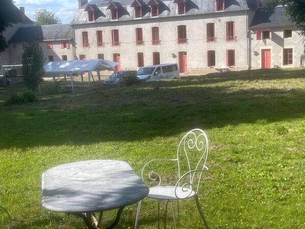 A grassy outdoor area with a metal table and chair, a building with red shutters in the background, and a parked van.