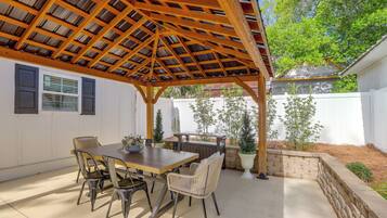 A covered patio area with a wooden pergola, a dining table with chairs, and potted plants.
