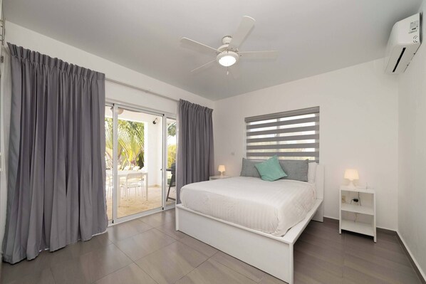 Ein Schlafzimmer mit einem Bett, einem Deckenventilator, einem Fenster mit Jalousien und einer Glastür, die zu einem Außenbereich führt.