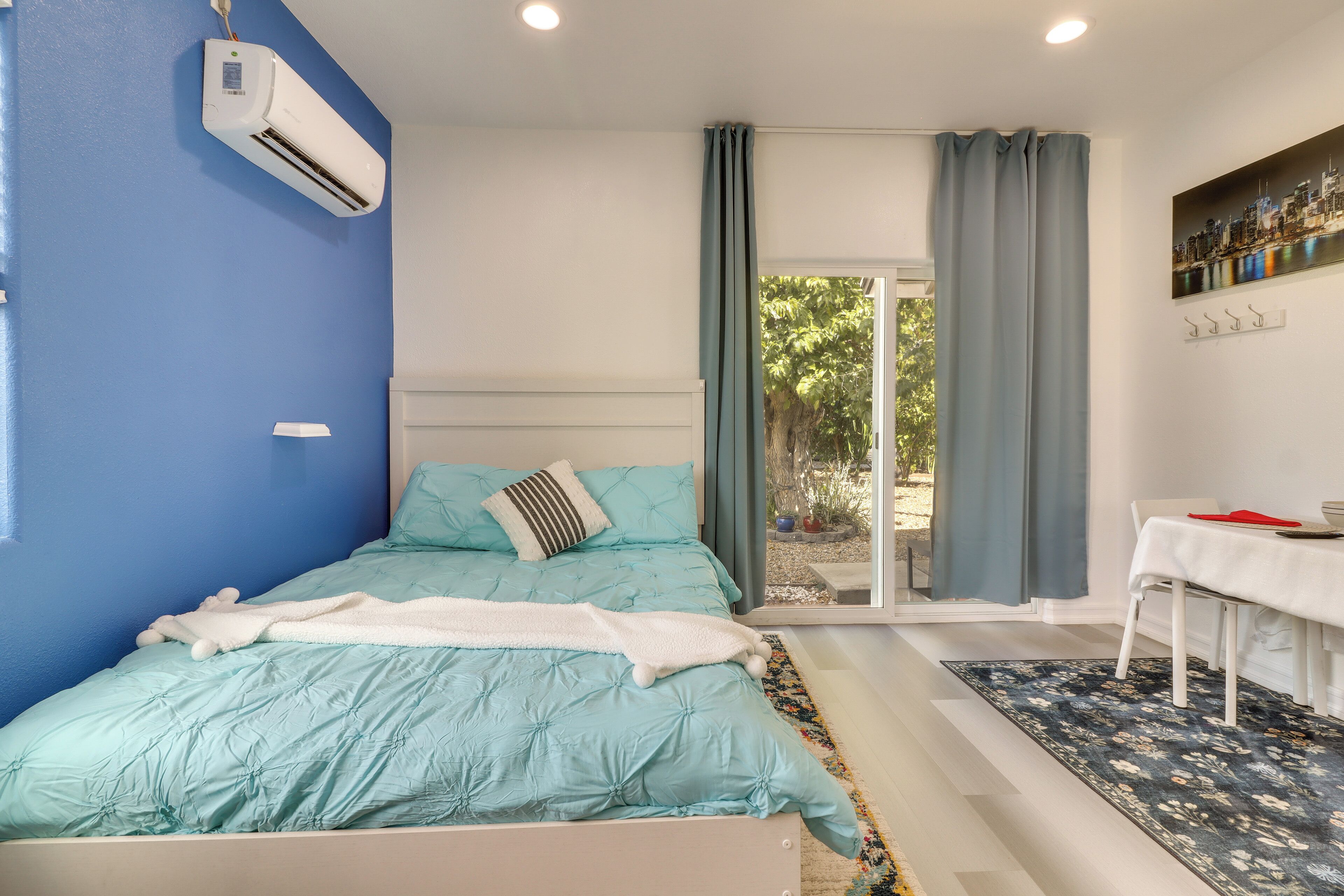 A bedroom with a bed, an air conditioning unit, a table with a red napkin, and a view of the outdoors through a glass door.