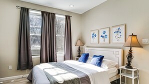 Ein Schlafzimmer mit einem Bett, zwei Nachttischlampen, einem Nachttisch, einem Fenster mit Vorhängen und zwei an der Wand befestigten Blumenbildern.